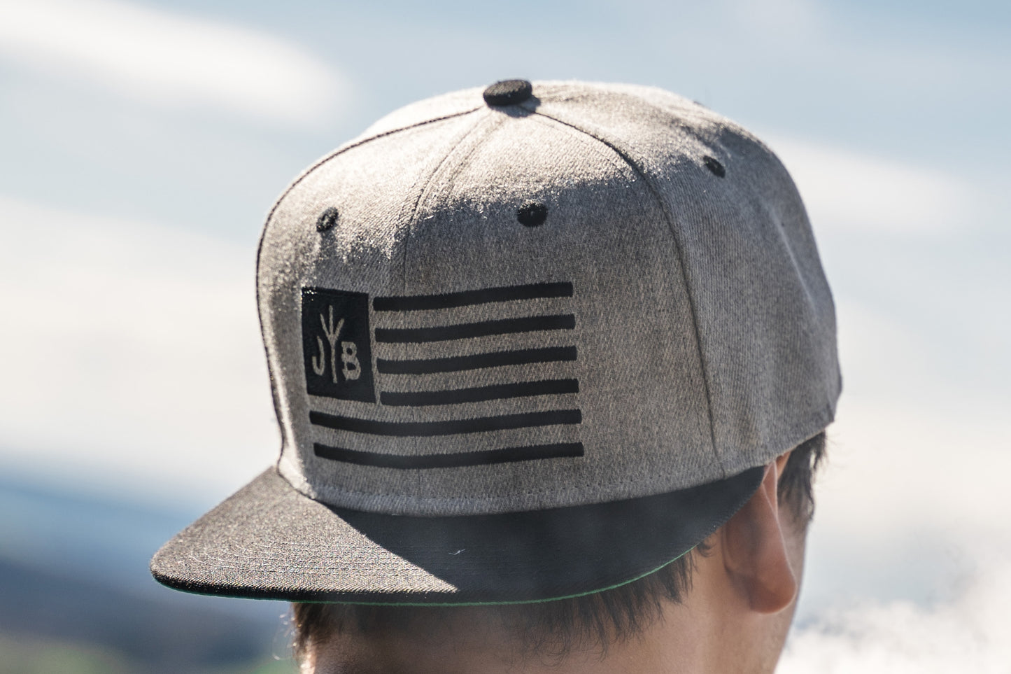 Snapback-Cap von Jumping Bird in Hellgrau-Schwarz mit gesticktem Flaggen-Design vorn, Logostick seitlich und grünem Schirm