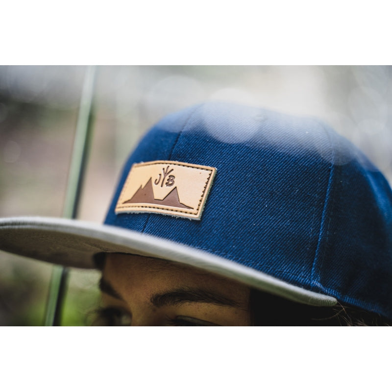 Snapback-Cap von Jumping Bird in Blau-Grau mit geprägtem Lederoptik-Patch vorn und seitlichem Logostick