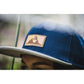 Snapback-Cap von Jumping Bird in Blau-Grau mit geprägtem Lederoptik-Patch vorn und seitlichem Logostick